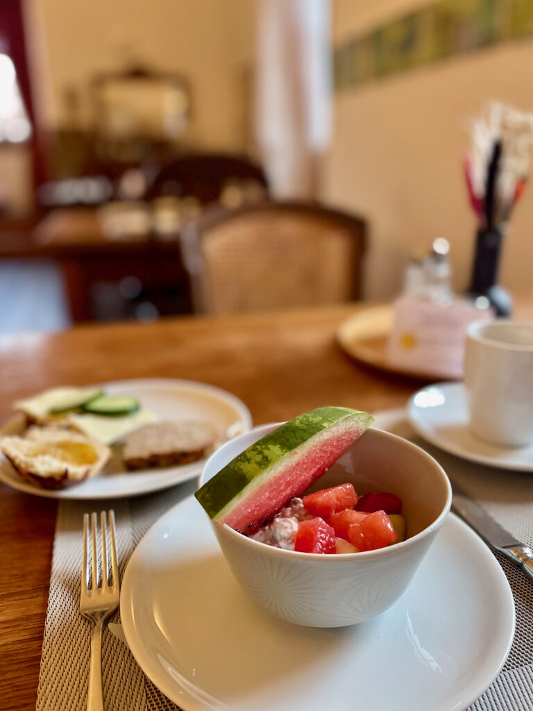 Fruehstueck im Resort Schloss Auerstedt. Am Fruehstueckstisch mit Muesli, Fruechten wie eine Scheibe Wassermelone, Ruehrei, Brot, Gurken und einer Tasse Kaffee. Im Hintergrund der Fruehstuecksraum.